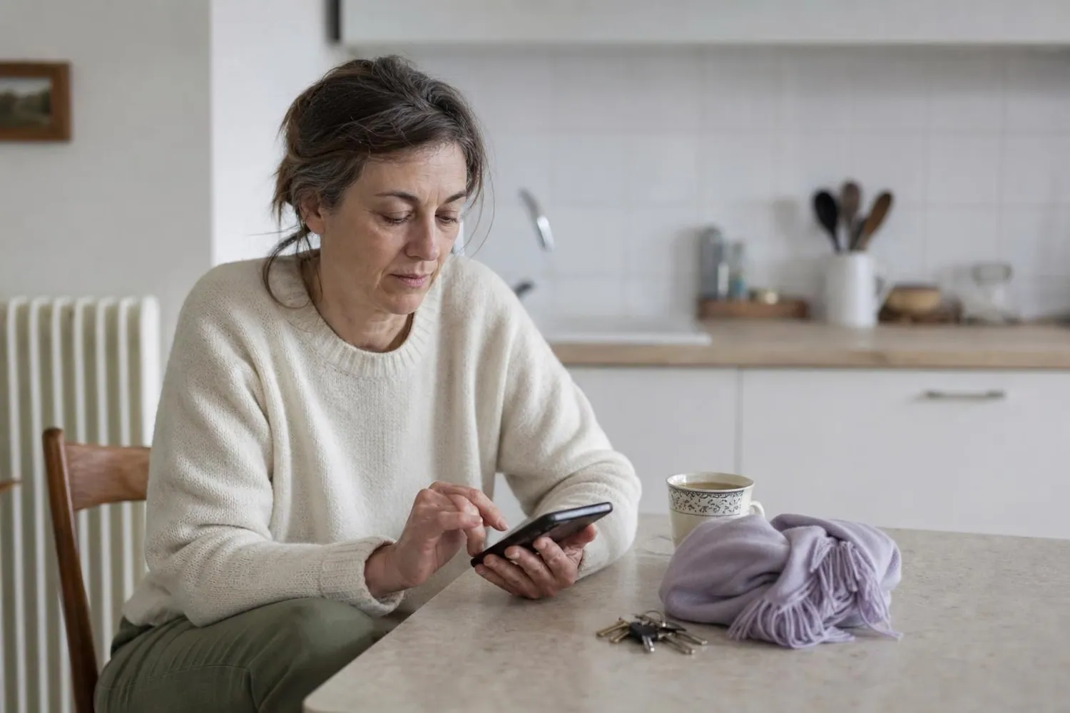 Femme d'une cinquantaine d'années assise dans sa cuisine, une tasse près d'elle, notant quelques mots sur son téléphone au petit matin
